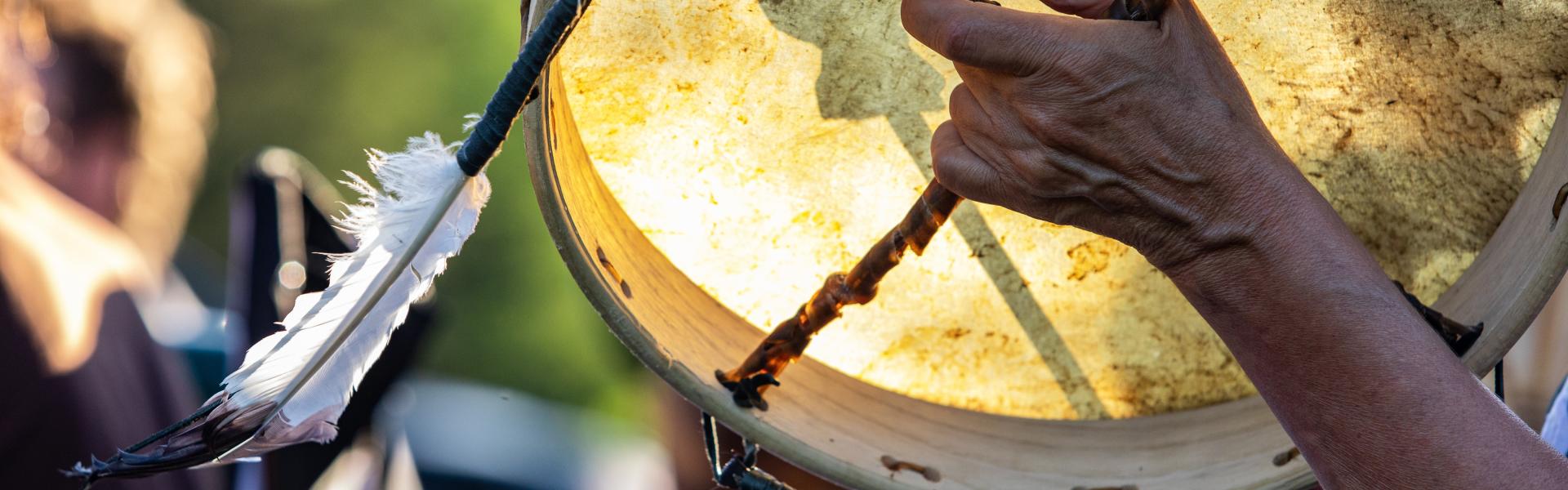 Traditionally constructed native drum is seen in the hands of a spiritual person with a sacred eagle feather during a park gig celebrating traditional music