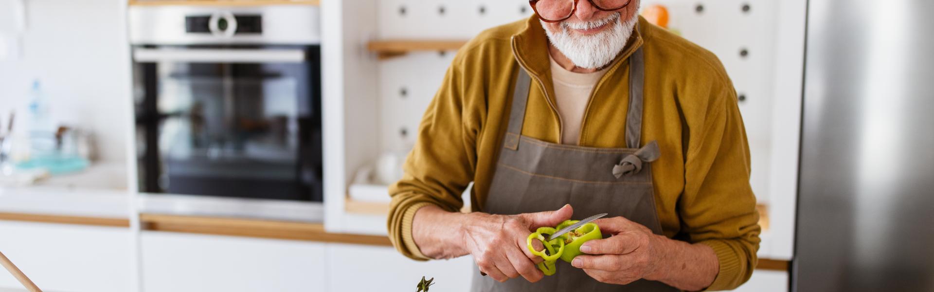 A older man cuts a green pepper while looking at a computer screen in the kitchen