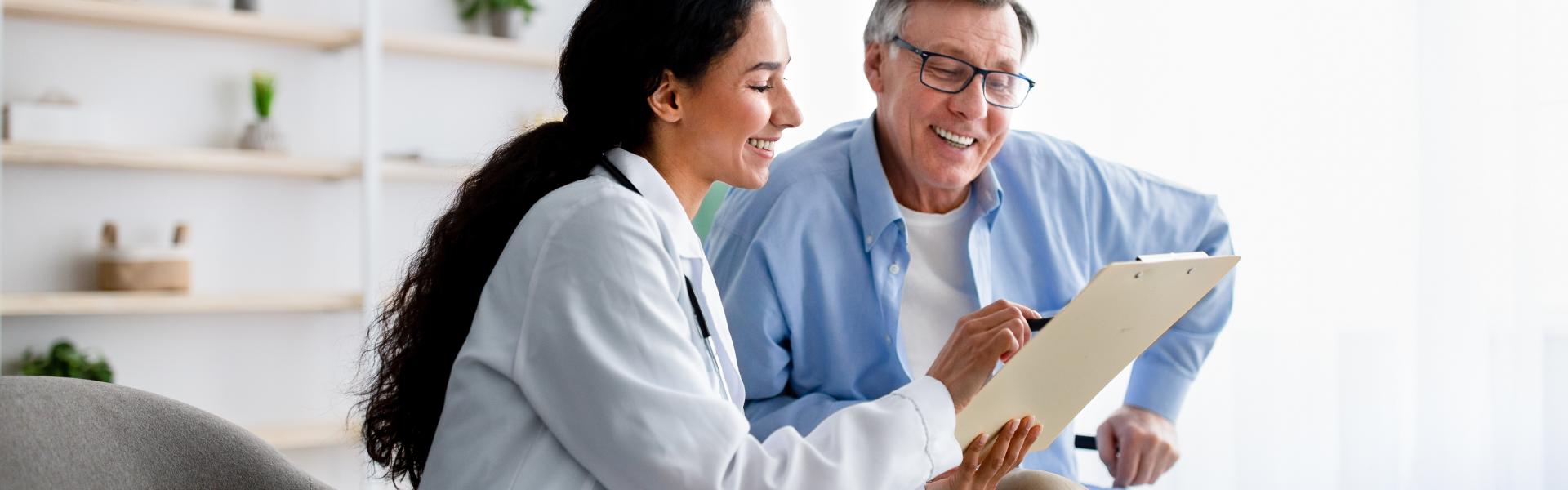 A doctor sits to review documents on a clipboard with an senior male patient in a wheelchair