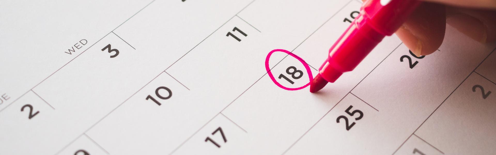 A white calendar on a table top with the 18th day of the month encircled in pink highlighter.