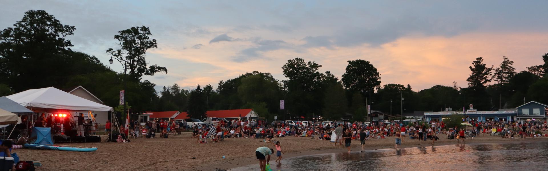 View of the entertainment tent and a large crowd on Balm Beach near sunset with the water and sand in the foreground.