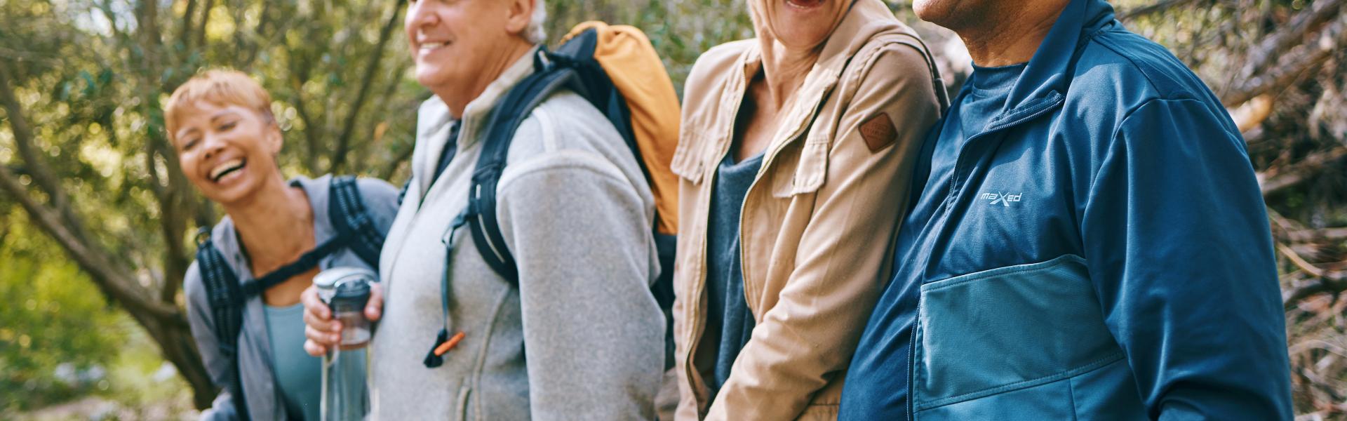 A group of four seniors dressed for fall weather, standing in a row in the forest and laughing.