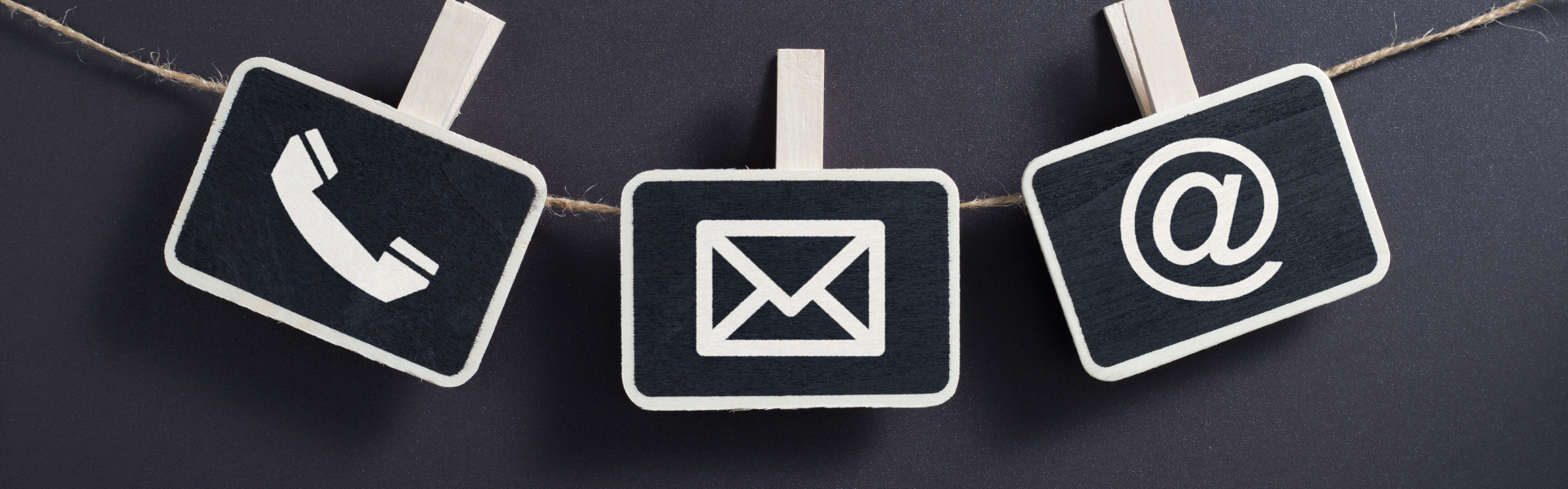 Three small wooden clothespins hanging from a string with a black background. On the first pin is a telephone symbol, on the second pin is an email symbol, and on the third pin is an at sign.