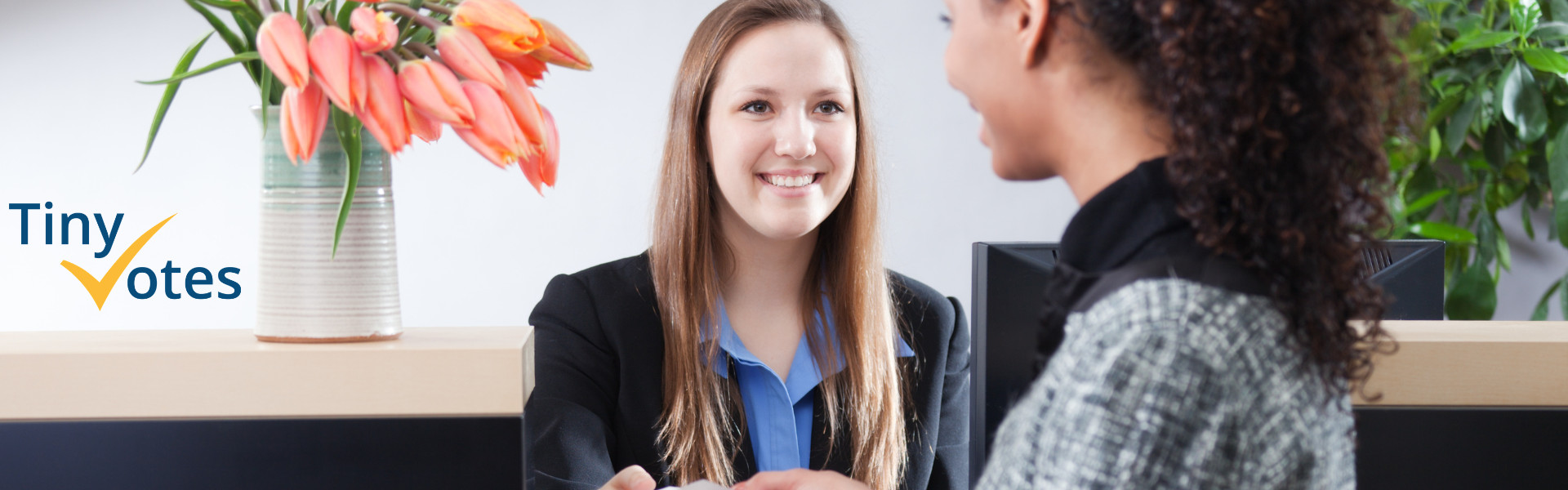 A woman smiling behind a service counter is assisting another woman as they exchange a piece of paper.