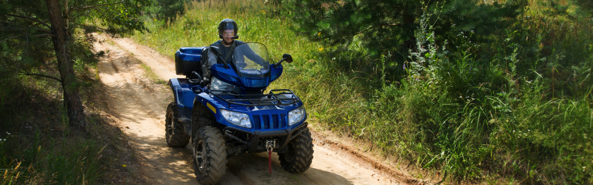 Rider wearing a helmet driving a blue ATV on a dirt pathway through a wooded area