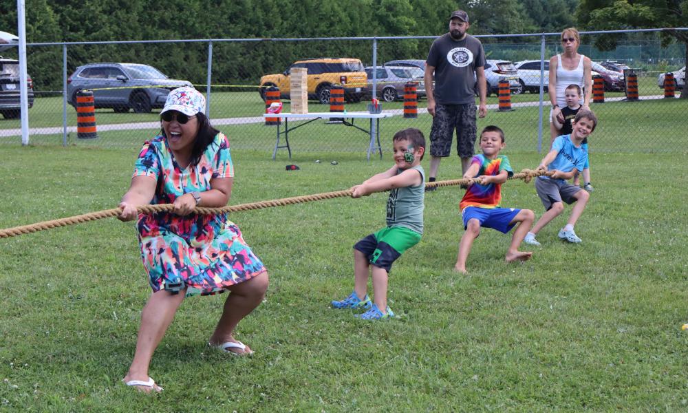 mother with several kids in bright clothing participating in a game of tug of war with other residents watching in the background