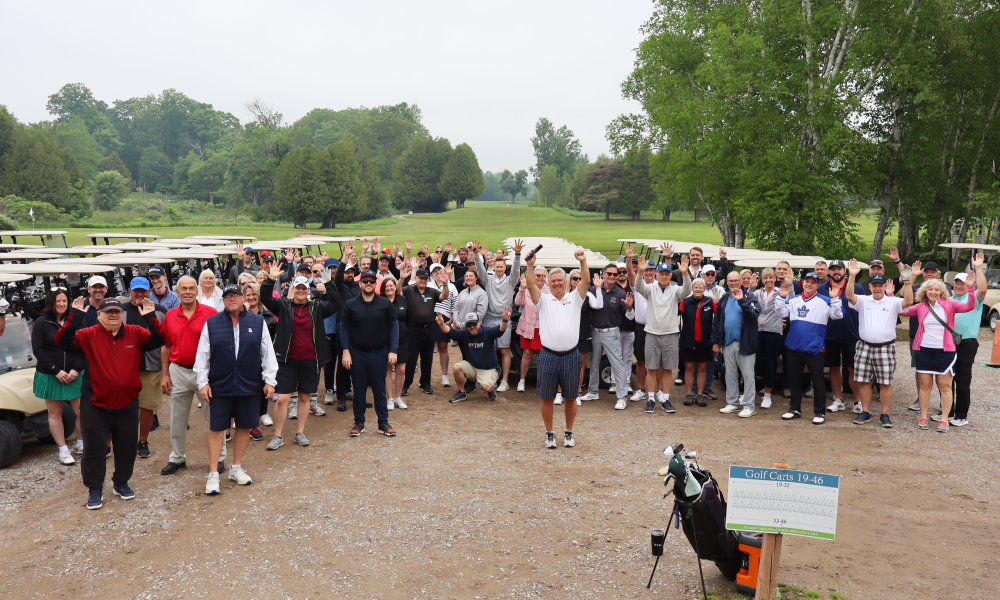 Large group photo of all of the golf participants standing and raising their arms up in cheer in front of the golf carts.
