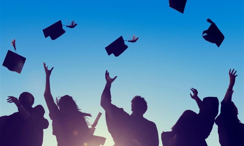 silhouette view of five students in graduation gowns throwing their caps in the air with the sun illuminating behind them and a bright blue sky