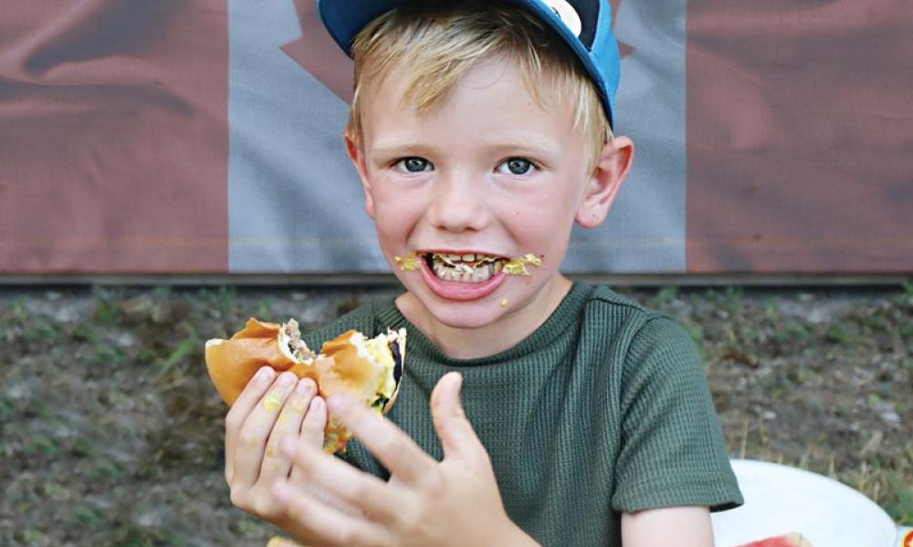 A boy wearing a baseball cap and eating a hamburger in front of a Canada Flag.