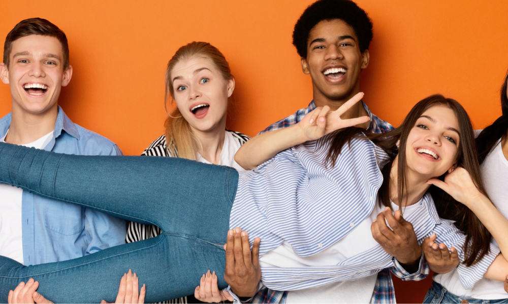 group of youth standing in front of a bright orange background. One of them is laying down and being held up by the others
