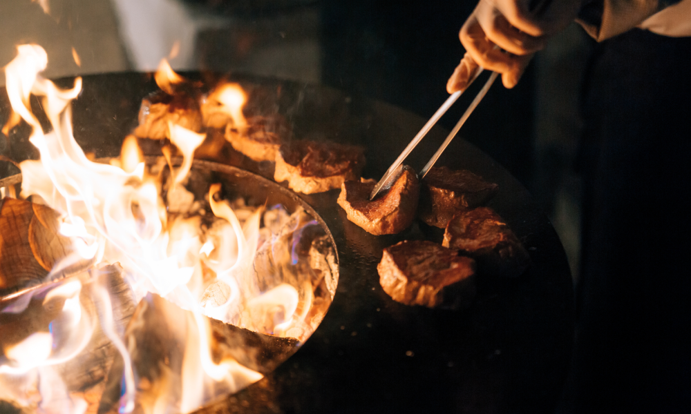 A person's hand holding tongs flipping pieces of meat on a fiery grill at night.