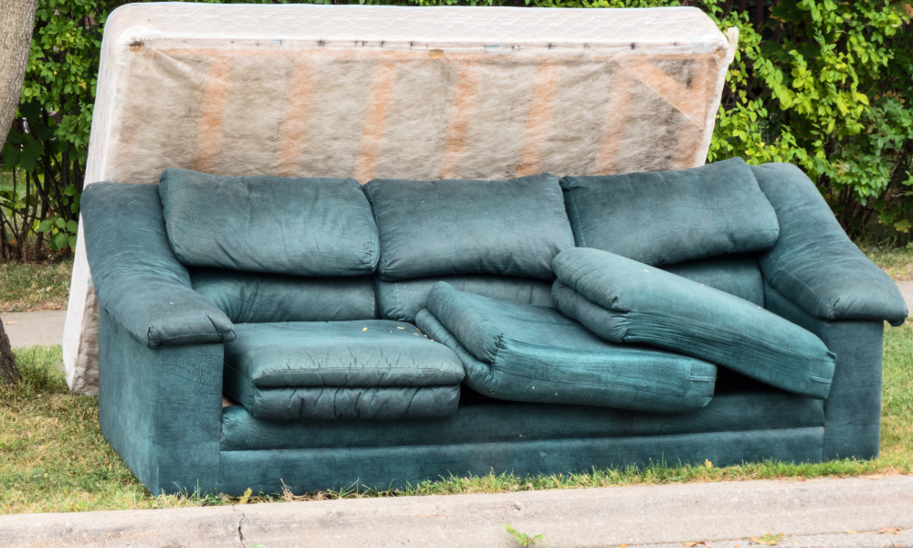 Green 3-seater couch with cushions not sitting in place and box spring leaning on the back of the couch on the side of the road