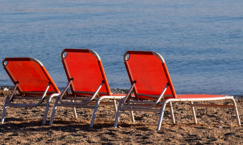 Three empty red and white lounge chairs lined up on a sandy beach near the water's edge.