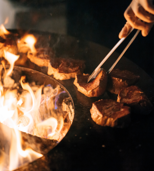 A person's hand holding tongs flipping pieces of meat on a fiery grill at night.