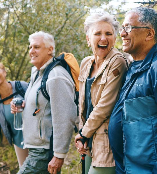 A group of four seniors dressed for fall weather, standing in a row in the forest and laughing.