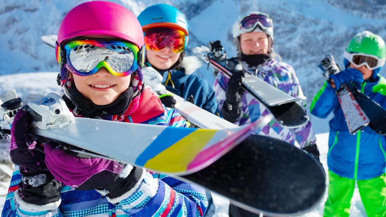 Happy smiling young girl in pink helmet and color goggles hold ski in her hands in a group of friends children on background over mountain