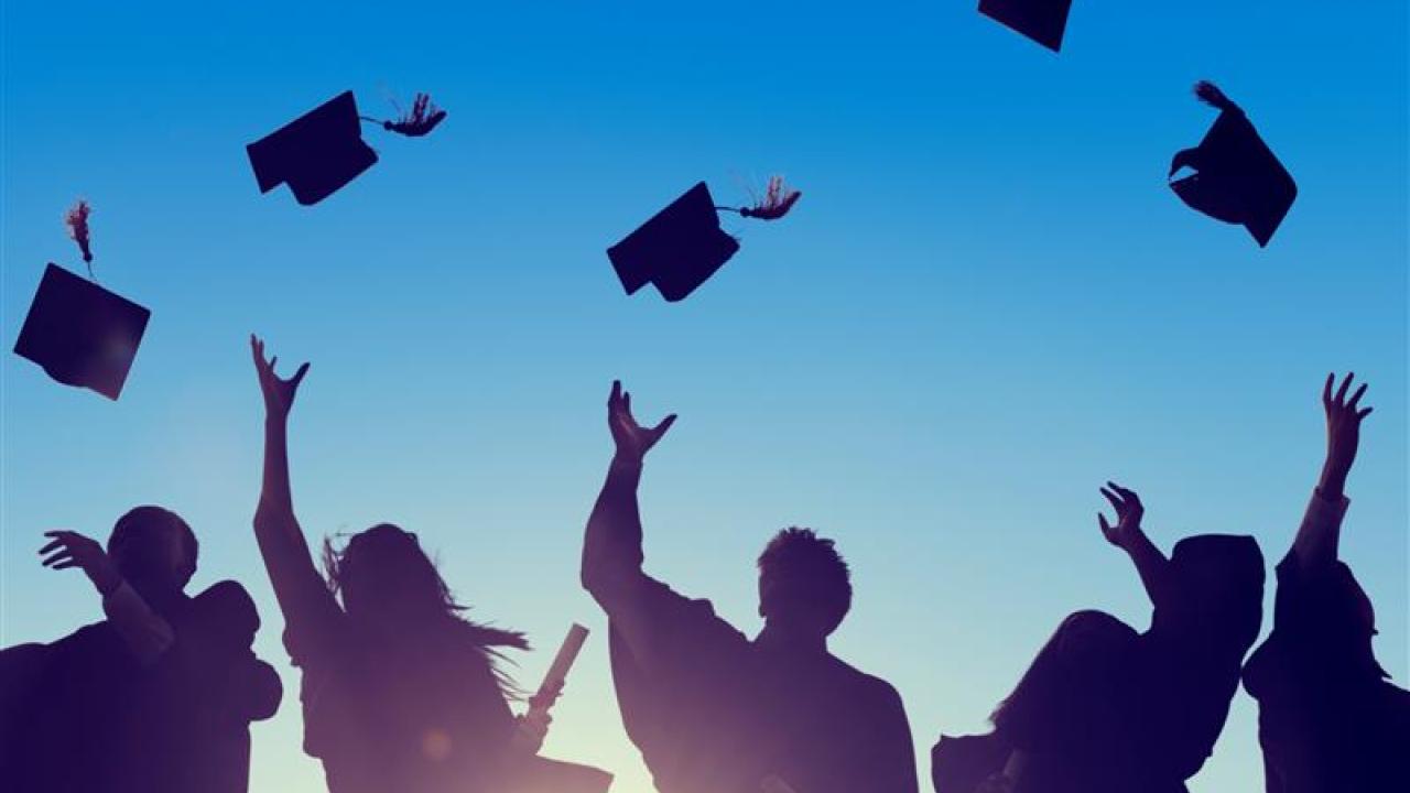 silhouette view of five students in graduation gowns throwing their caps in the air with the sun illuminating behind them and a bright blue sky