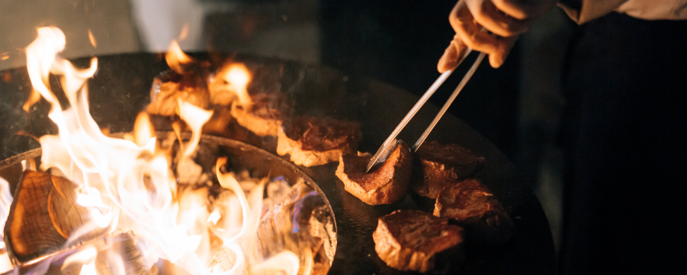 A hand holding cooking tongs, cooking steaks on an open campfire