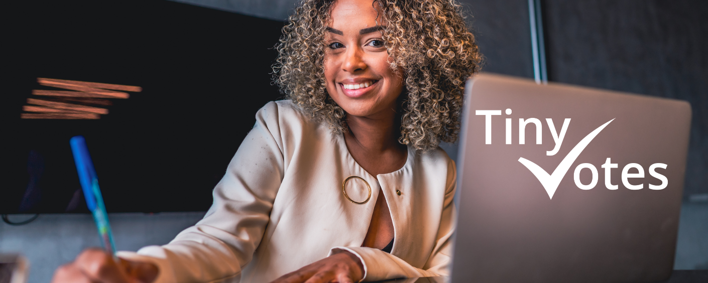woman with curly hair sitting at a desk with a laptop and paper and pen smiling at the camera