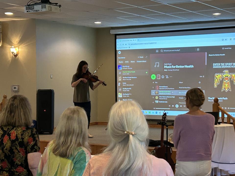 Ladies standing around a table at the Senior Symposium listening to a music therapist play violin