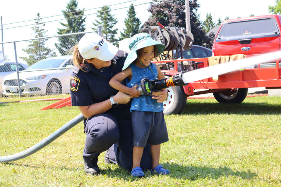 female firefighter in their blue uniform and Tiny baseball cap helping a young boy wearing a blue outfit and big blue hat, smiling, use the fire hose
