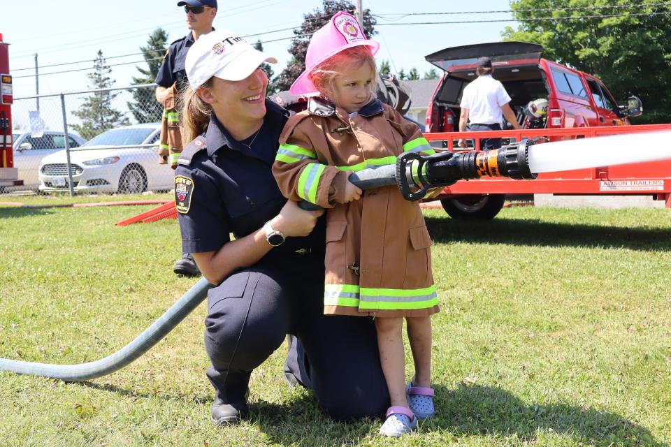 young girl dressed in a child-sized firefighter jacket that comes down to their knees and a pink firefighter helmet with a female firefighter spraying water from the fire hose