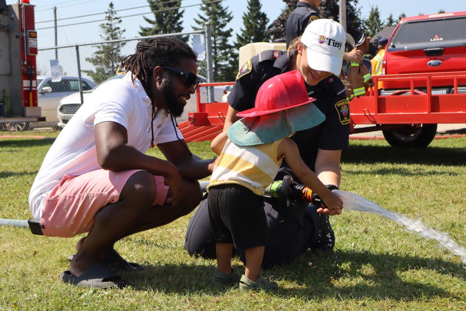female firefighter encouraging a toddler to touch the light pressure water coming from the fire hose with the child's dad watching