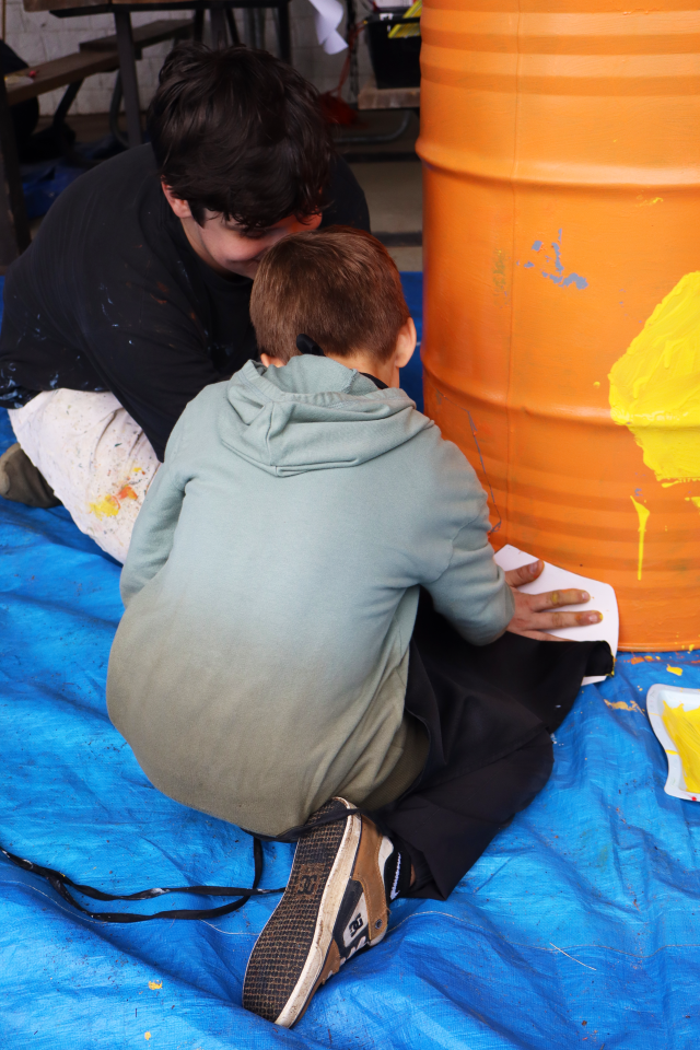 one of the local artists helping a young student paint a yellow hexagon on the orange barrel