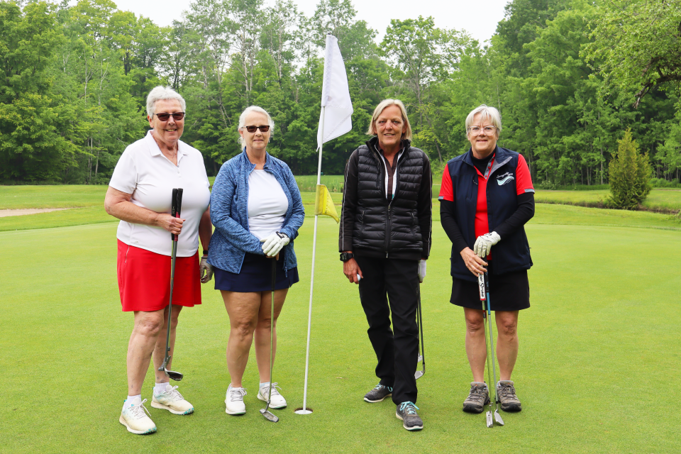 Golf foursome of all women standing next to a white flag, holding their golf clubs