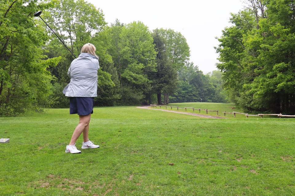 Woman as their golf swing is all the way through. While hard to see, the golf ball can be spotted high in the sky at the tops of the trees on the longest drive hole.