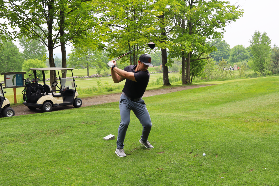 Man mid-swing focused on their golf ball with golf carts and trees in the background.