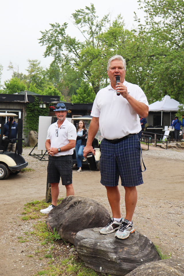 Mayor David Evans standing on top of a rock with a microphone addressing the golf participants with Deputy Mayor Miskimins standing beside him. Golf carts and the club house can be seen in the background.