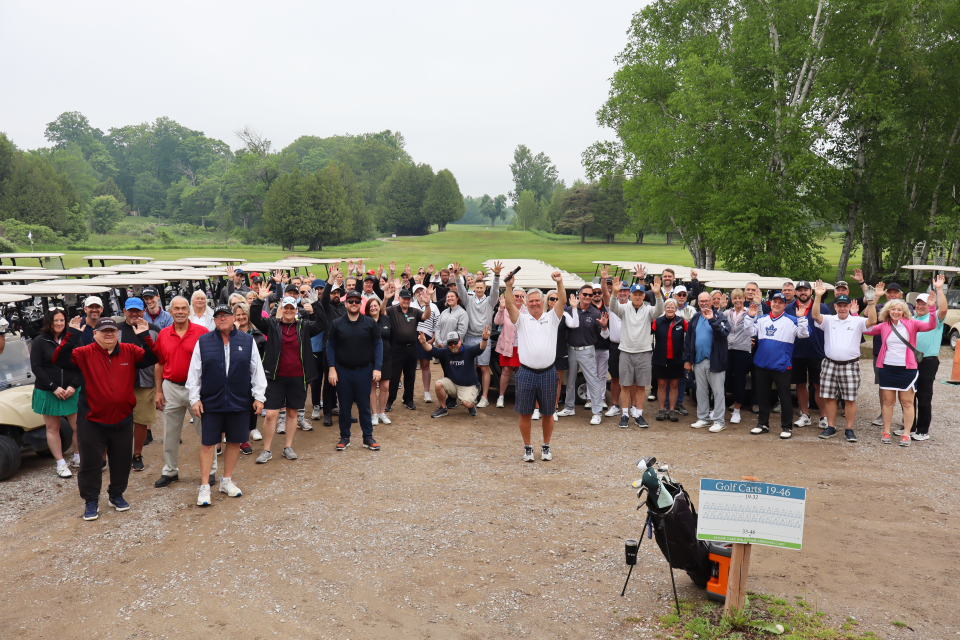 Large group photo of all of the golf participants standing and raising their arms up in cheer in front of the golf carts.