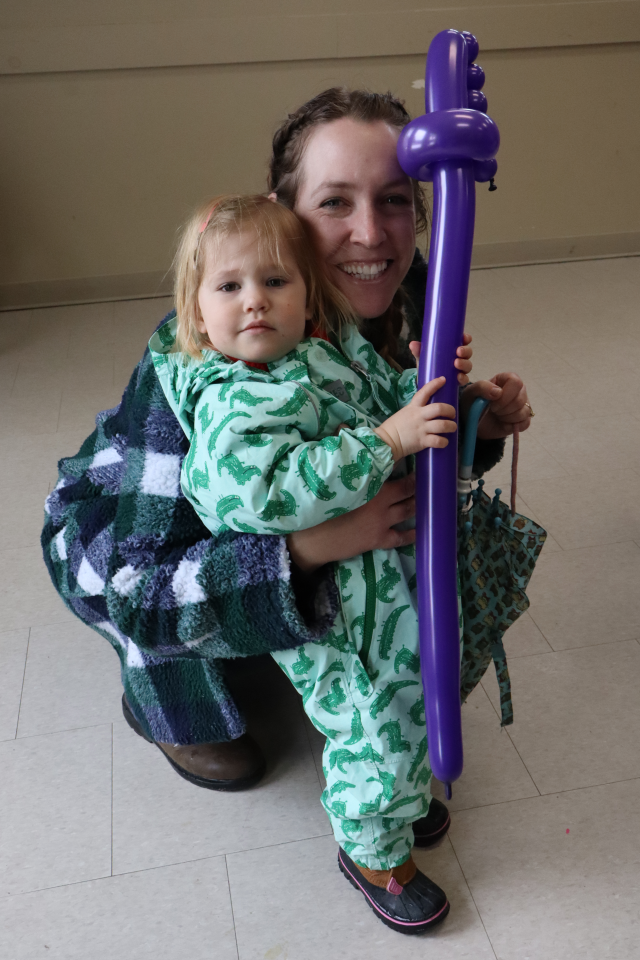 Young mom and child dressed in a bright green patterned snow suit, holding a purple balloon twisted to look like a sword