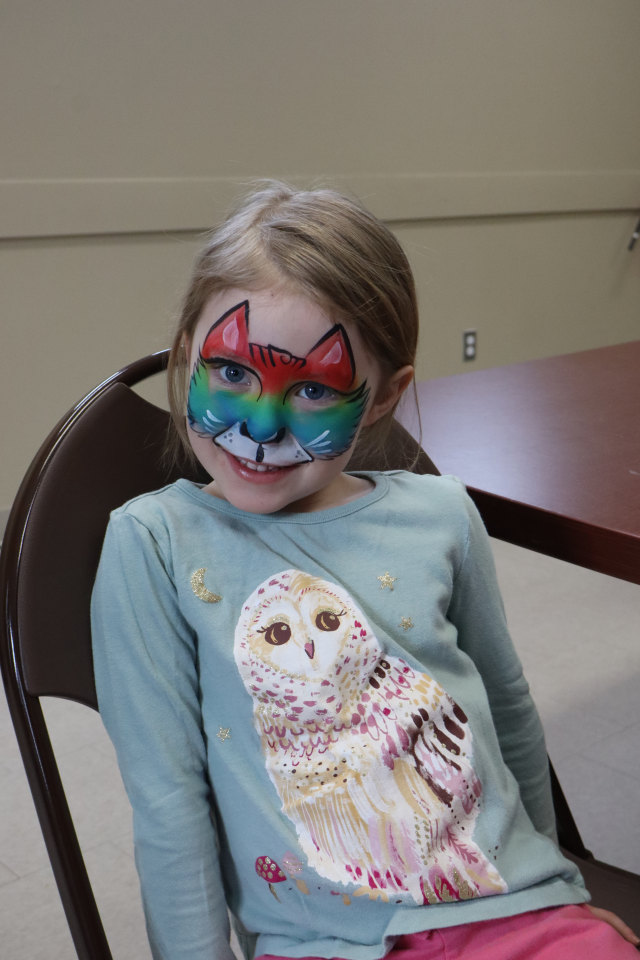 Girl with a bright rainbow cat facepaint design sitting in a chair wearing a blue shirt with a large owl