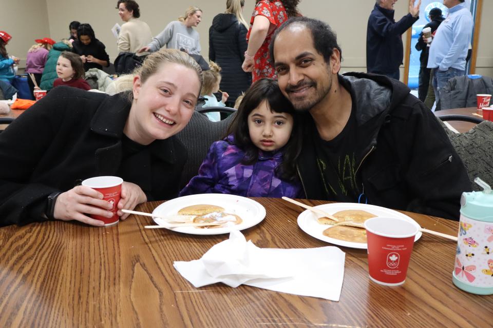 Young family with a small child in a bright purple coat sitting down at a table to enjoy pancakes and hot chocolate