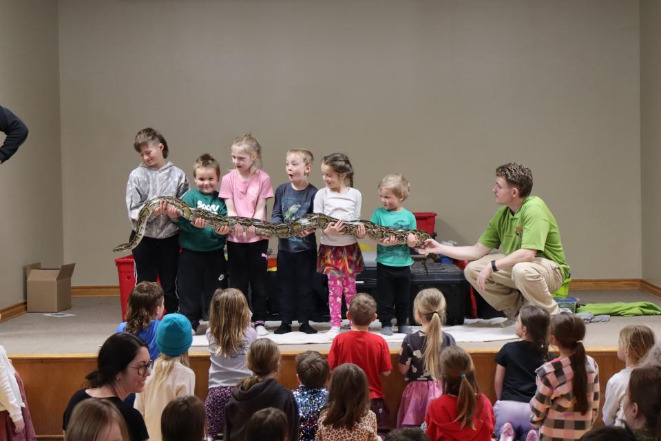 several children standing in a row holding a large snake with the help of Zoo to You staff