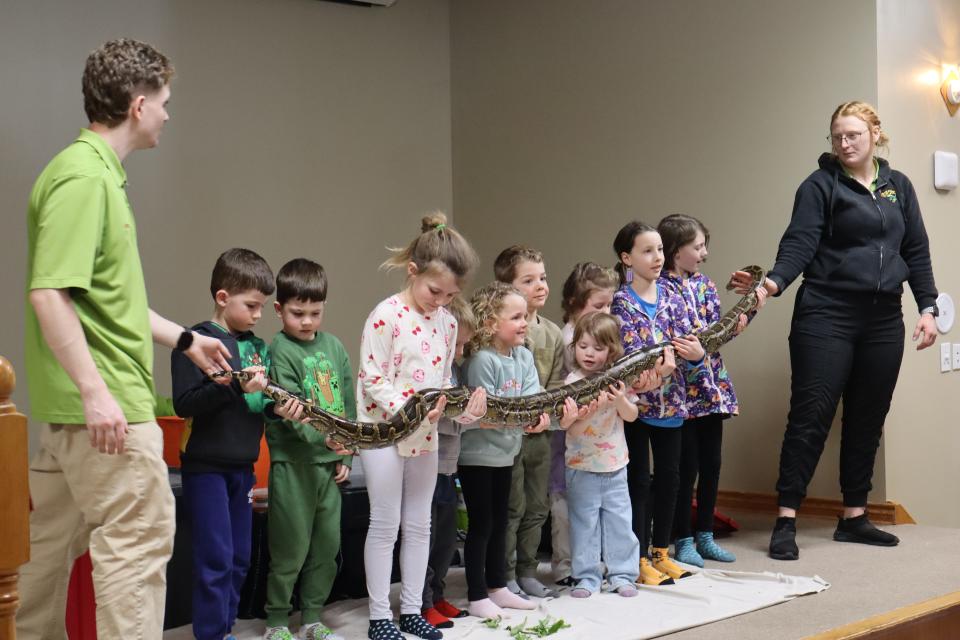 Group of children standing in a row holding a large snake with the help of Zoo to You staff