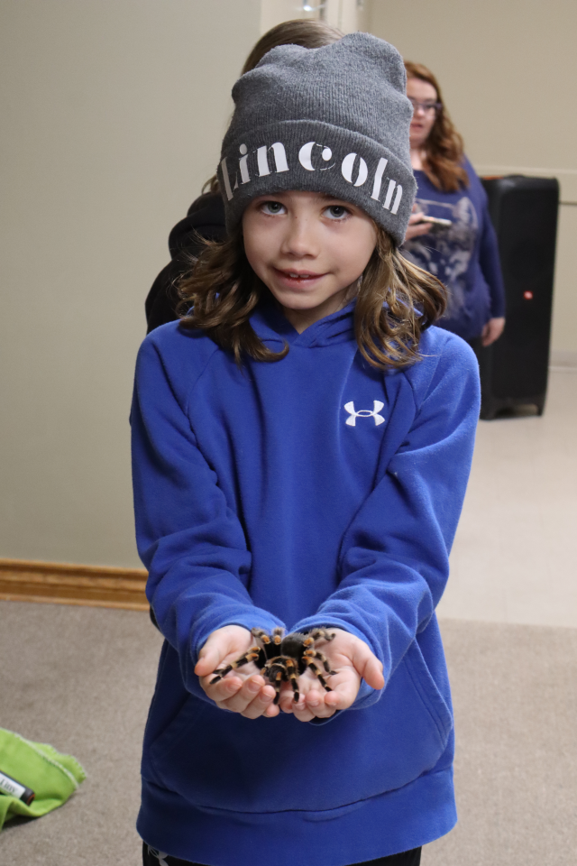 Child in a blue sweatshirt and grey hat standing on stage holding a tarantula