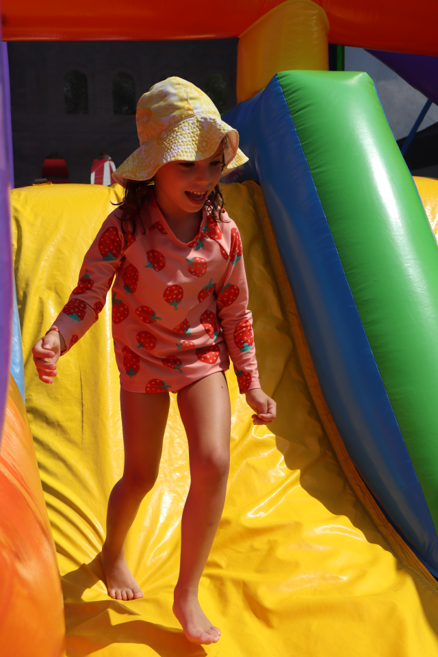 Child wearing a bright pink outfit and hat jumping down a brightly coloured slide of a bouncy castle.
