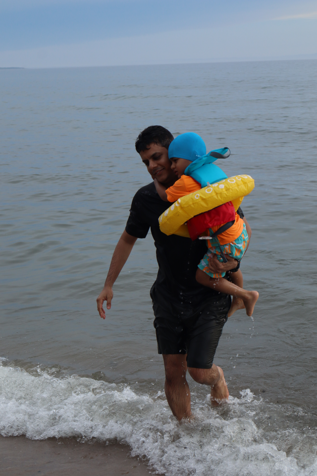 Father carrying their son out of the water at Balm Beach, the child is wearing a brightly coloured swimsuit, life jacket, and inner tube.