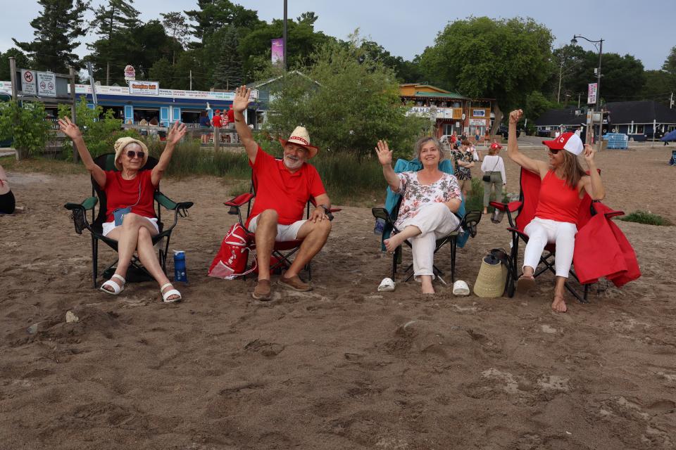 Group of four adults waving to the camera while seated in comfortable lawn chairs in the sand.