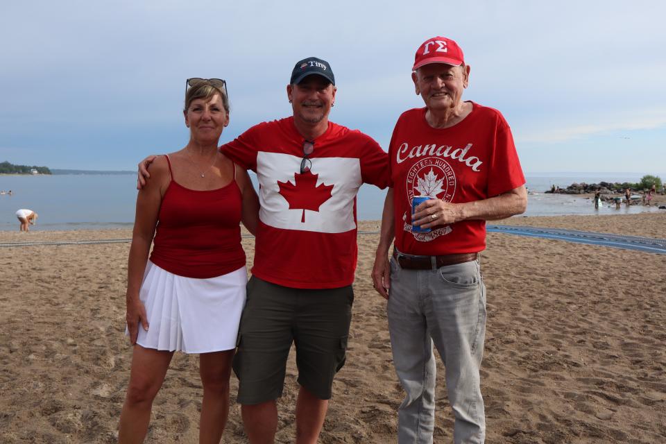 Group of three adults posing on the beach with the open water in the background wearing their red and white colours, with Deputy Mayor Sean Miskimins in the middle.