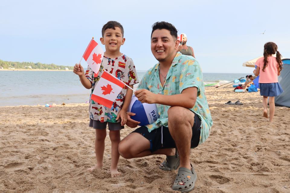 Father and son holding Canadian flags wearing brightly coloured clothing on the sand.