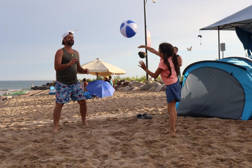Man and girl playing with a blue and white beach ball on the sand with tents in the background.