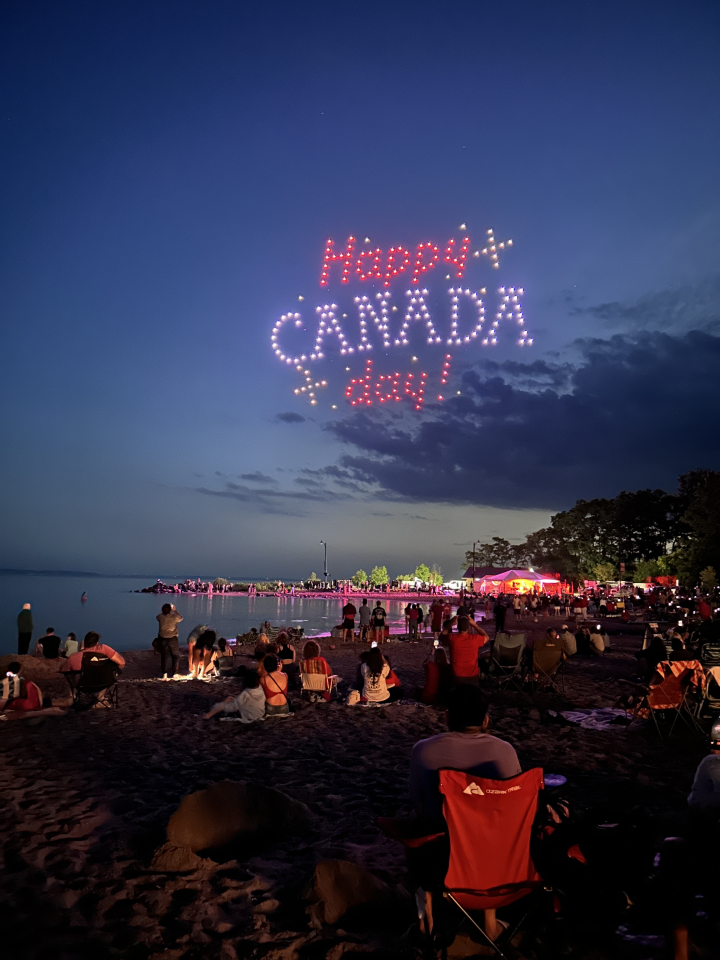 View of the drone show with Happy Canada Day written in the sky with a view behind the crowd and lights emanating from the stage. Photo credit Melanie Robitaille @mwords_photo