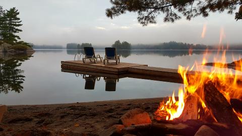 campfire on the beach with a dock and muskoka chairs sitting on the dock facing away from the camera