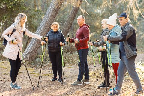 Serious female sports trainer with group of people talking, explaining posture for Nordic walking with sticks in forest 