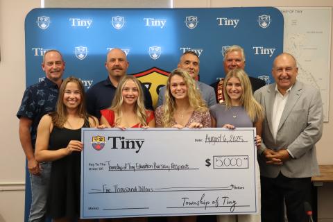 Council with 4 of the 5 2025 education bursary winners posing with a large Tiny branded novelty cheque in front of the Tiny photo wall