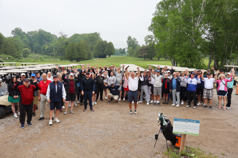 Large group photo of all of the golf participants standing and raising their arms up in cheer in front of the golf carts.